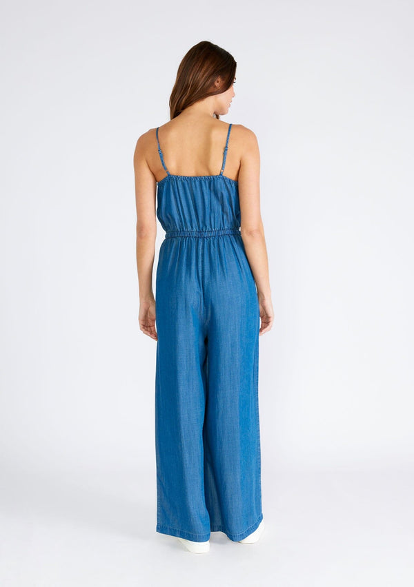 [Color: Vintage Wash] A back facing image of a brunette model wearing a classic denim blue sleeveless jumpsuit crafted from Tencel. With adjustable spaghetti straps, a scoop neckline, a self covered button front top, an adjustable drawstring tie waist, side pockets, and a long wide leg. 