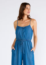 [Color: Vintage Wash] A close up front facing image of a brunette model wearing a classic denim blue sleeveless jumpsuit crafted from Tencel. With adjustable spaghetti straps, a scoop neckline, a self covered button front top, an adjustable drawstring tie waist, side pockets, and a long wide leg. 