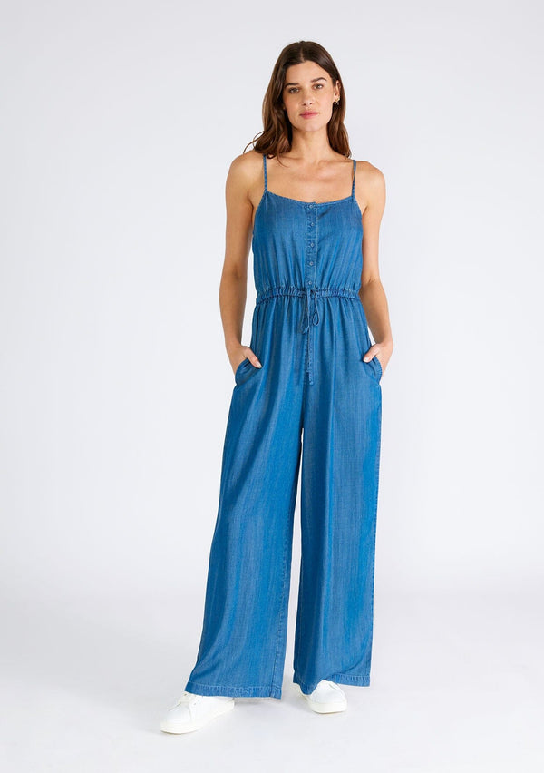 [Color: Vintage Wash] A front facing image of a brunette model wearing a classic denim blue sleeveless jumpsuit crafted from Tencel. With adjustable spaghetti straps, a scoop neckline, a self covered button front top, an adjustable drawstring tie waist, side pockets, and a long wide leg. 