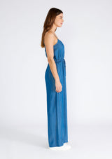 [Color: Vintage Wash] A side facing image of a brunette model wearing a classic denim blue sleeveless jumpsuit crafted from Tencel. With adjustable spaghetti straps, a scoop neckline, a self covered button front top, an adjustable drawstring tie waist, side pockets, and a long wide leg. 