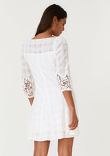 [Color: Off White] A back facing image of a brunette model wearing a white cotton bohemian mini dress in a textured dobby plaid fabric. With a flared paneled mini skirt, a scooped neckline, three quarter length sleeves with a crochet detail, and a relaxed fit. 
