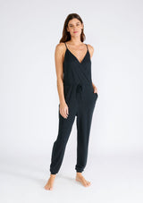 [Color: Black] A front facing image of a brunette model wearing a black ribbed knit sleeveless jumpsuit. With spaghetti straps, a surplice v neckline, side pockets, and a drawstring tie waist. 