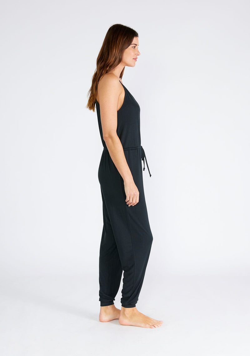 [Color: Black] A side facing image of a brunette model wearing a black ribbed knit sleeveless jumpsuit. With spaghetti straps, a surplice v neckline, side pockets, and a drawstring tie waist. 