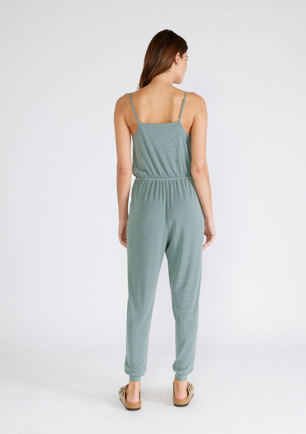 [Color: Slate Green] A back facing image of a brunette model wearing a seafoam green ribbed knit sleeveless jumpsuit. With spaghetti straps, a surplice v neckline, side pockets, and a drawstring tie waist. 