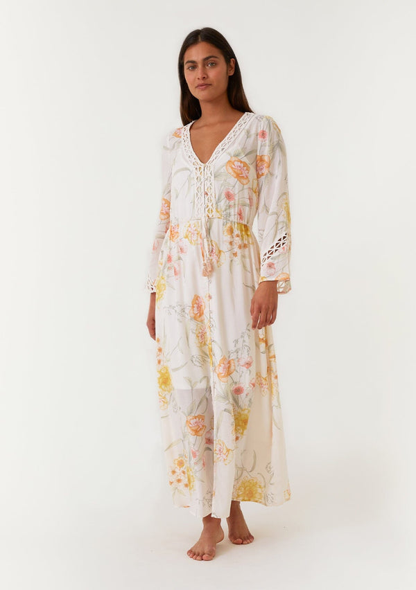 [Color: Vanilla/Coral] A front facing image of a brunette model wearing a dreamy bohemian maxi dress designed in an ivory and coral pink floral print. With long bell sleeves, a v neckline, a self covered button front, a long flowy skirt with front slit, lace trim, and an adjustable drawstring waist with tassel ties. 