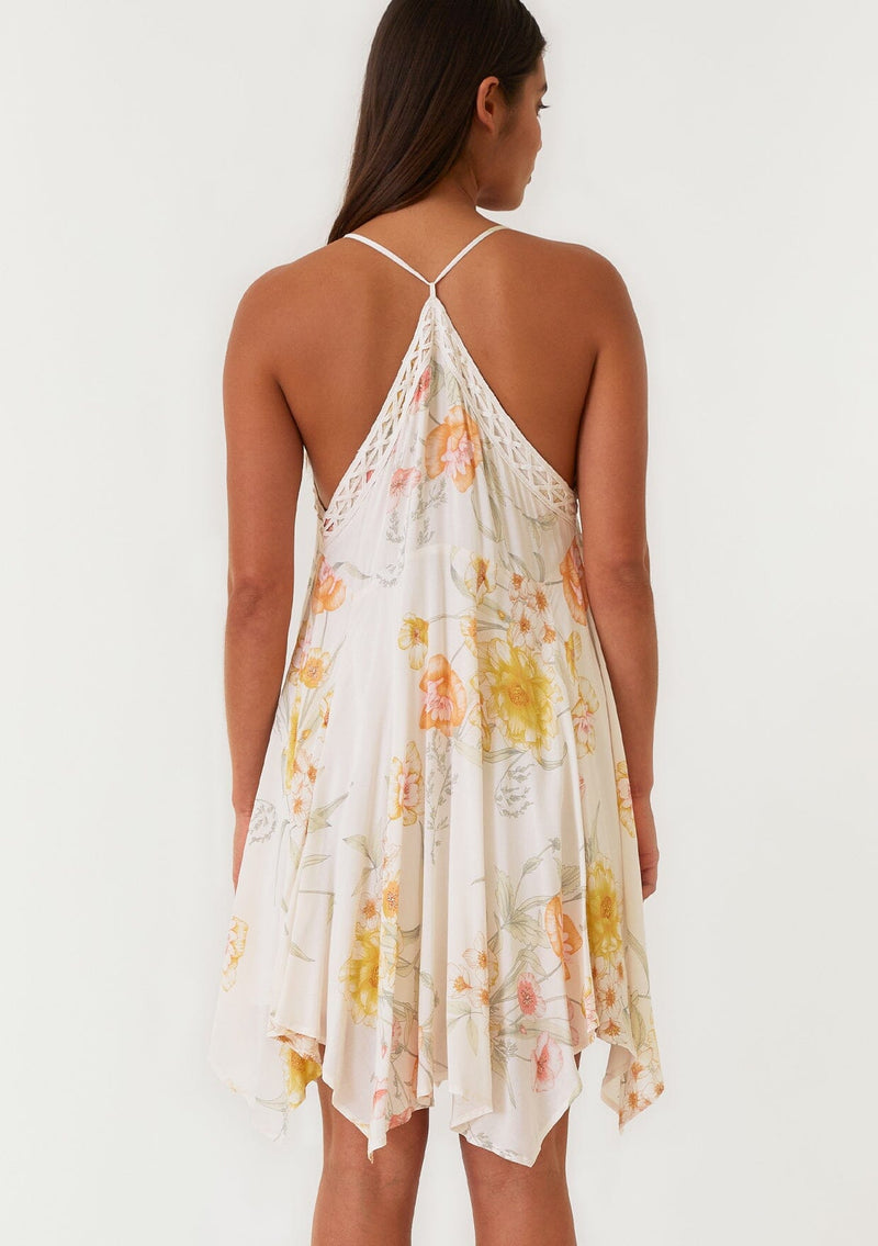 [Color: Vanilla/Coral] A back facing image of a brunette model wearing a flowy bohemian mini dress in an ivory and coral floral print. With spaghetti straps, a v neckline, a racer back detail, an asymmetric hemline, and lace trim.