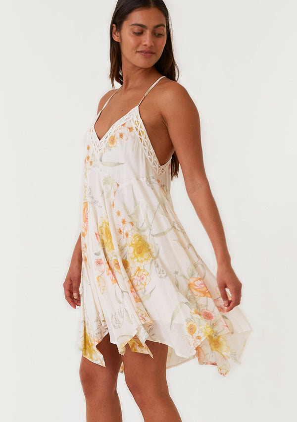 [Color: Vanilla/Coral] A side facing image of a brunette model wearing a flowy bohemian mini dress in an ivory and coral floral print. With spaghetti straps, a v neckline, a racer back detail, an asymmetric hemline, and lace trim.