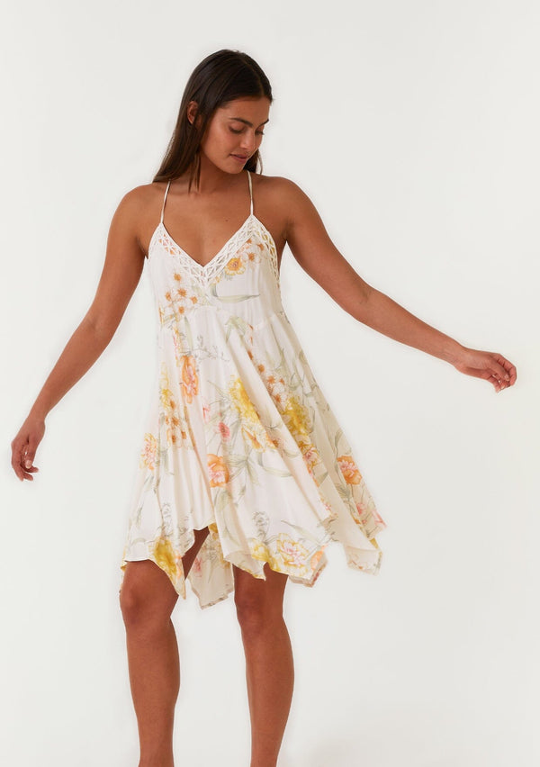 [Color: Vanilla/Coral] A full body front facing image of a brunette model wearing a flowy bohemian mini dress in an ivory and coral floral print. With spaghetti straps, a v neckline, a racer back detail, an asymmetric hemline, and lace trim.