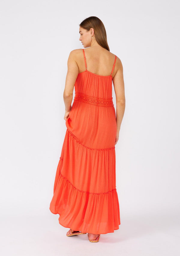 [Color: Coral] A back facing image of a brunette model wearing a bright coral red sleeveless maxi dress. With adjustable spaghetti straps, a straight neckline, crochet trim, a tassel tie drawstring waist, and a ruffle trimmed tiered skirt. 