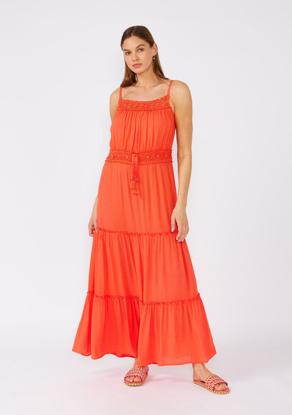 [Color: Coral] A front facing image of a brunette model wearing a bright coral red sleeveless maxi dress. With adjustable spaghetti straps, a straight neckline, crochet trim, a tassel tie drawstring waist, and a ruffle trimmed tiered skirt. 