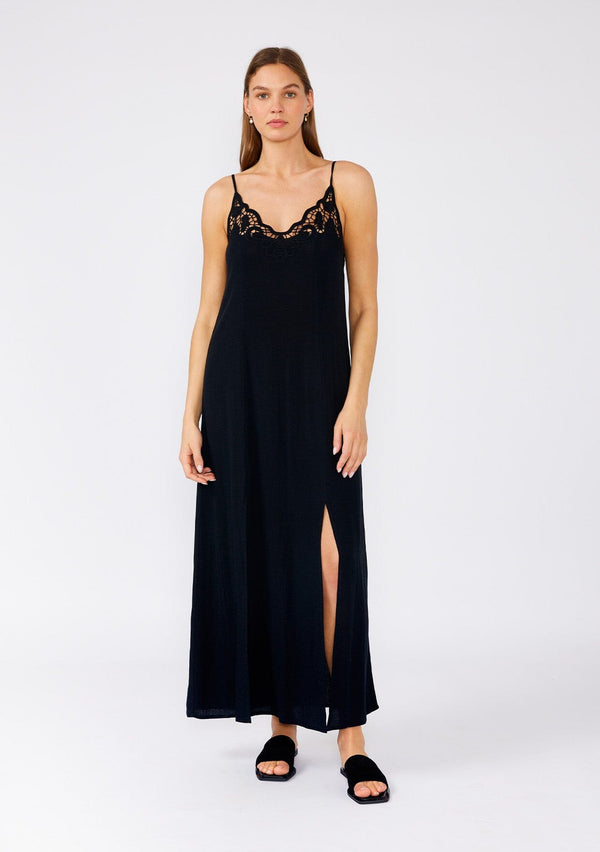[Color: Black] A front facing image of a brunette model wearing a classic black maxi slip dress with lace detail along the neckline. With adjustable spaghetti straps, a scooped neckline, and a long flowy skirt with side slits. 