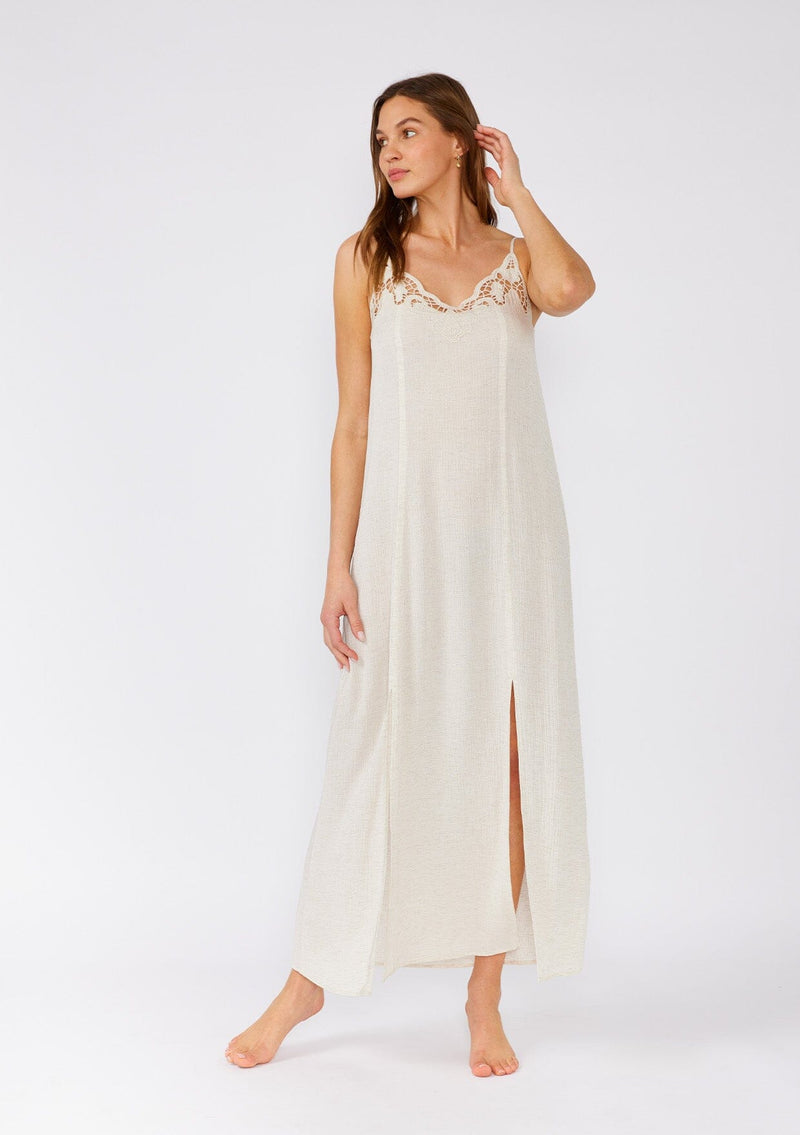 [Color: Natural] A front facing image of a brunette model wearing a classic off white maxi slip dress with lace detail along the neckline. With adjustable spaghetti straps, a scooped neckline, and a long flowy skirt with side slits. 