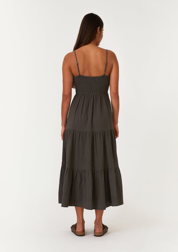 [Color: Lagoon] A back facing image of a brunette model wearing a sleeveless army green mid length spring dress. With adjustable spaghetti straps, a scoop neckline, a tiered skirt, and a half smocked elastic bodice at the back. 