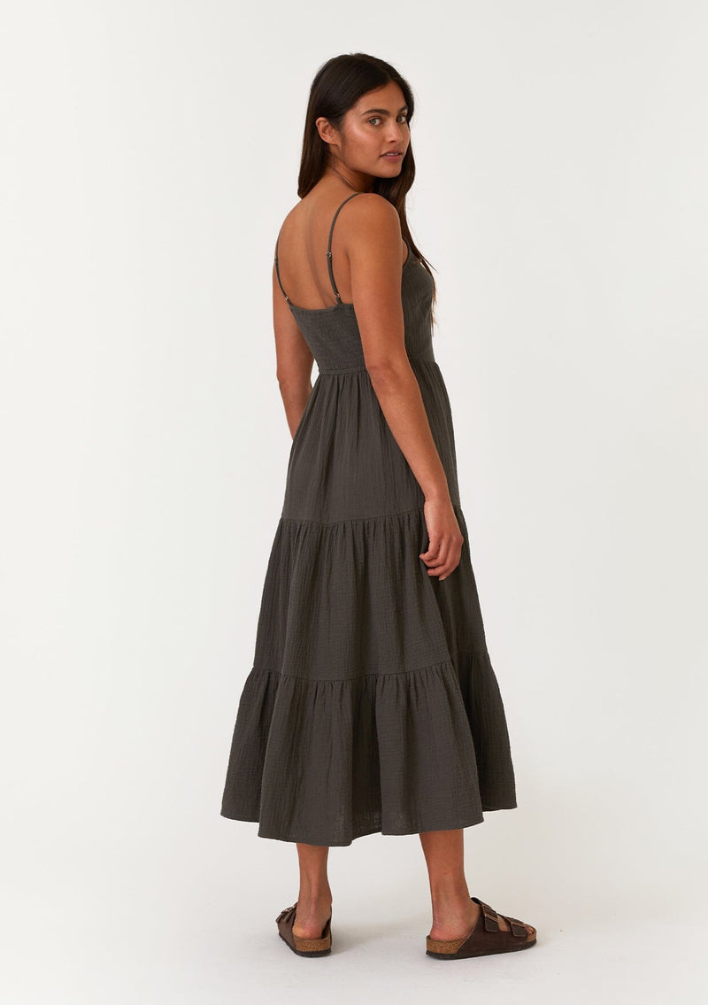 [Color: Lagoon] A full body back facing image of a brunette model wearing a sleeveless army green mid length spring dress. With adjustable spaghetti straps, a scoop neckline, a tiered skirt, and a half smocked elastic bodice at the back. 