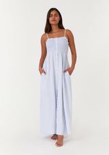 [Color: Dusty Blue] A front facing image of a brunette model wearing a light blue bohemian maxi dress made with soft cotton gauze. With adjustable spaghetti straps, a smocked bodice, a long flowy skirt, a self covered button front, side pockets, an empire waist, and a straight neckline with ruffle trim. 