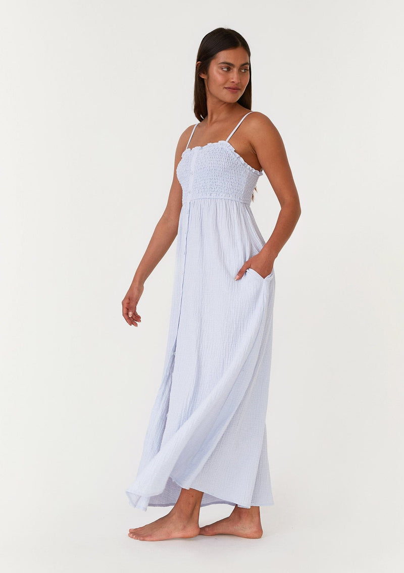 [Color: Dusty Blue] A side facing image of a brunette model wearing a light blue bohemian maxi dress made with soft cotton gauze. With adjustable spaghetti straps, a smocked bodice, a long flowy skirt, a self covered button front, side pockets, an empire waist, and a straight neckline with ruffle trim. 