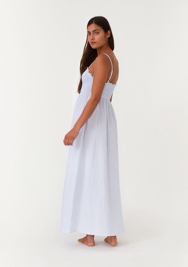 [Color: Dusty Blue] A back facing image of a brunette model wearing a light blue bohemian maxi dress made with soft cotton gauze. With adjustable spaghetti straps, a smocked bodice, a long flowy skirt, a self covered button front, side pockets, an empire waist, and a straight neckline with ruffle trim. 
