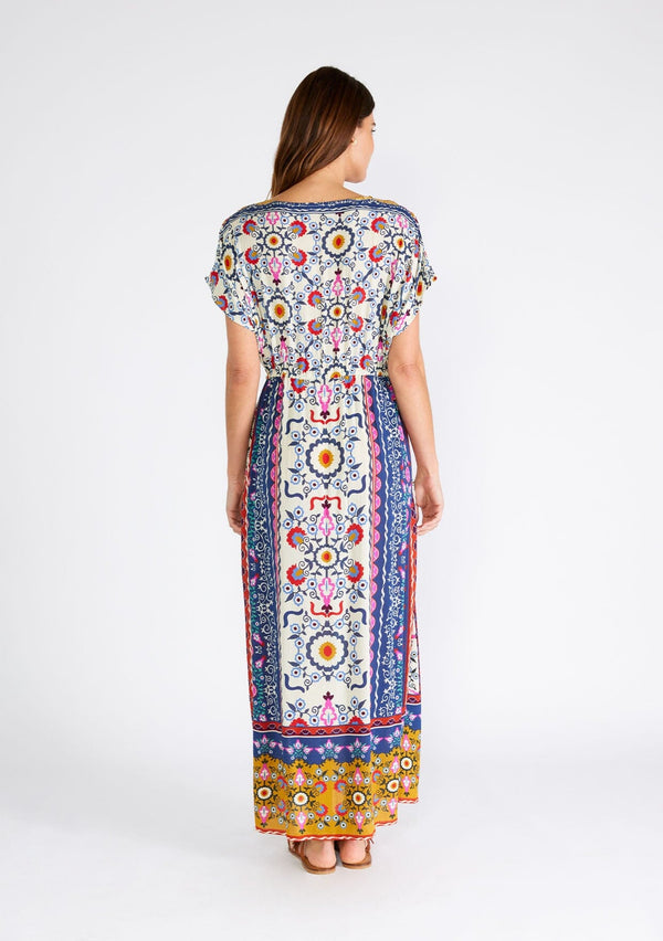 [Color: Natural/Mustard] A back facing image of a brunette model wearing a brightly colorful bohemian maxi dress in a mixed floral print. With short dolman sleeves, a v neckline, a self covered button front top, an adjustable drawstring tie waist, and a long flowy skirt with side slits. 