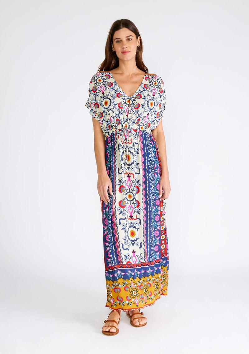 [Color: Natural/Mustard] A full body front facing image of a brunette model wearing a brightly colorful bohemian maxi dress in a mixed floral print. With short dolman sleeves, a v neckline, a self covered button front top, an adjustable drawstring tie waist, and a long flowy skirt with side slits. 