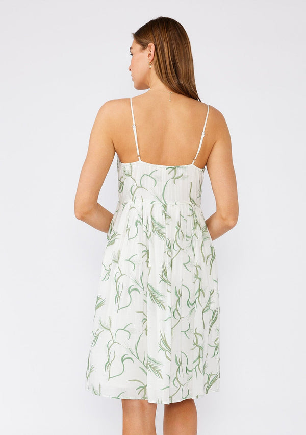 [Color: Off White/Green] A back facing image of a brunette model wearing a sleeveless cotton bohemian mini dress designed in a white and green embroidery. With spaghetti straps, an empire waist, a scooped neckline, side pockets, and a flowy silhouette. 