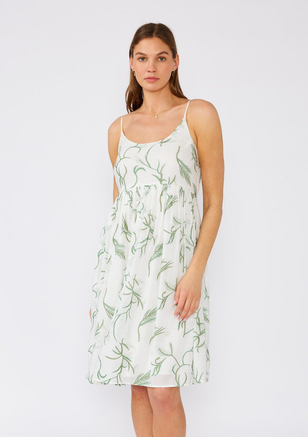 [Color: Off White/Green] A front facing image of a brunette model wearing a sleeveless cotton bohemian mini dress designed in a white and green embroidery. With spaghetti straps, an empire waist, a scooped neckline, side pockets, and a flowy silhouette. 