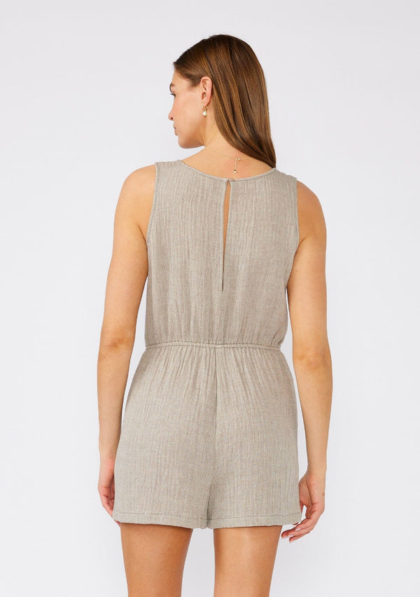 [Color: Taupe] A back facing image of a brunette model wearing a sleeveless light brown cotton short romper. With a scooped neckline, side pockets, and a back keyhole with single button loop closure. 