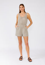 [Color: Taupe] A full body front facing image of a brunette model wearing a sleeveless light brown cotton short romper. With a scooped neckline, side pockets, and a back keyhole with single button loop closure. 
