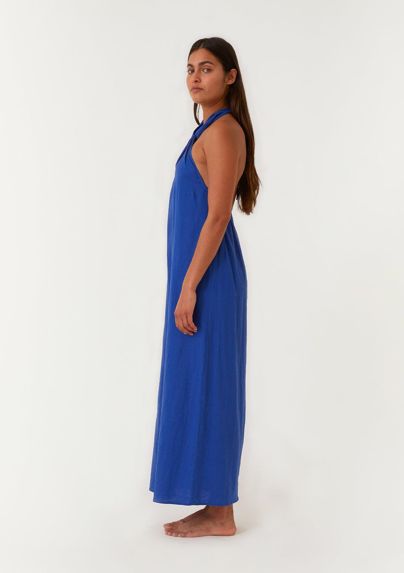 [Color: Cobalt] A side facing image of a brunette model wearing a bright cobalt blue halter maxi dress. With a twist front halter neckline, adjustable tie, front slit, and sexy open back. Perfect for the beach or the pool. 