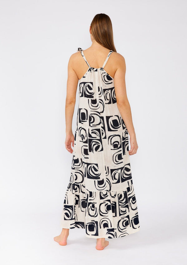 [Color: Ivory/Black] A back facing image of a brunette model wearing a flowy sleeveless bohemian maxi dress designed in an ivory and black print. With a tiered skirt and a drawstring neckline with adjustable tie. 