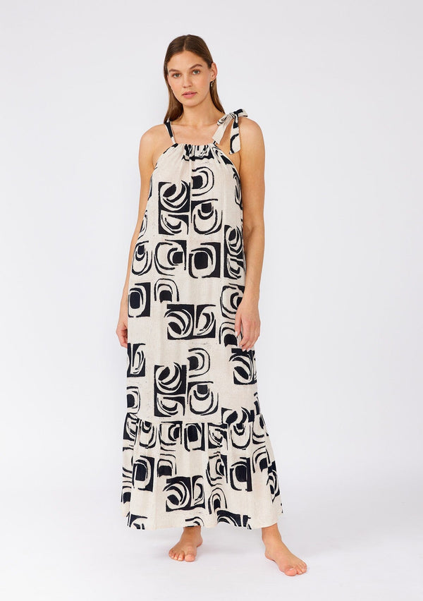 [Color: Ivory/Black] A front facing image of a brunette model wearing a flowy sleeveless bohemian maxi dress designed in an ivory and black print. With a tiered skirt and a drawstring neckline with adjustable tie. 