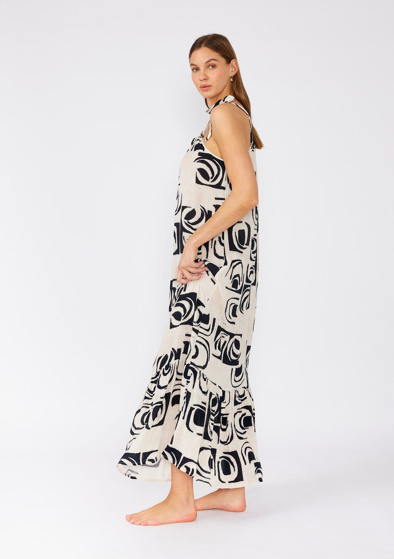 [Color: Ivory/Black] A side facing image of a brunette model wearing a flowy sleeveless bohemian maxi dress designed in an ivory and black print. With a tiered skirt and a drawstring neckline with adjustable tie. 