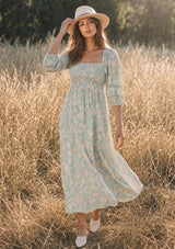 A woman stands in a sunlit field wearing a floral midi dress with three-quarter sleeves, a square neckline, and a smocked bust. She is also wearing a wide-brim hat and white mules.