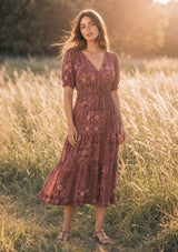 A woman wearing a burgundy floral midi dress standing in a sunlit grassy field.