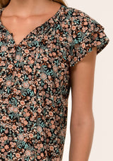 [Color: Black/Teal] A close up front facing image of a blonde model wearing a bohemian fall blouse in a black and teal floral print. With a self covered button front, double flutter short cap sleeves, and neck ties. 
