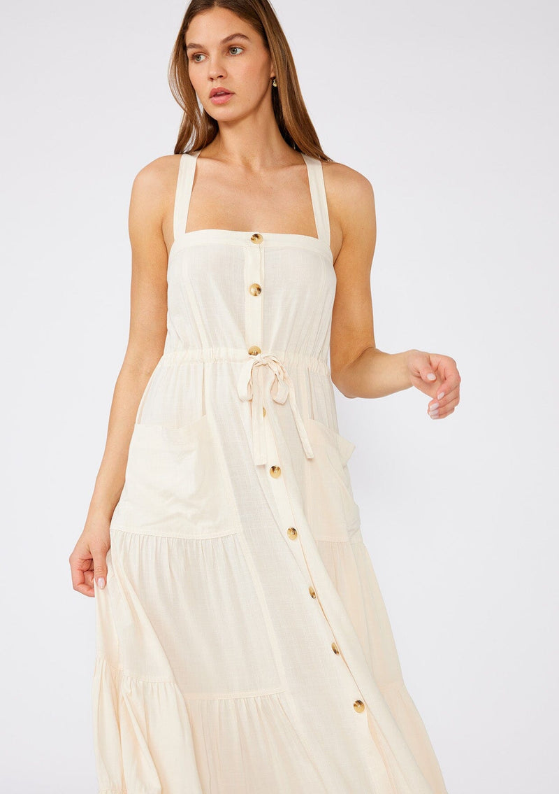 [Color: Vanilla] A close up front facing image of a brunette model wearing an off white linen blend sleeveless maxi dress. With a billowy tiered skirt, a large tortoise shell button front, an adjustable drawstring waist, and a cross back strap detail.