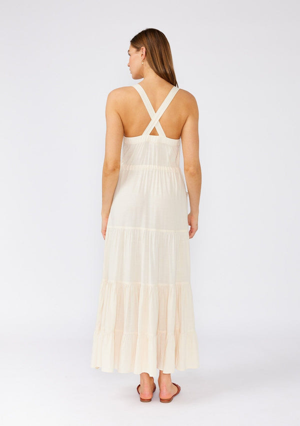 [Color: Vanilla] A back facing image of a brunette model wearing an off white linen blend sleeveless maxi dress. With a billowy tiered skirt, a large tortoise shell button front, an adjustable drawstring waist, and a cross back strap detail.