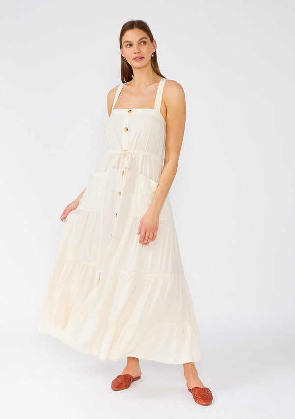[Color: Vanilla] A front facing image of a brunette model wearing an off white linen blend sleeveless maxi dress. With a billowy tiered skirt, a large tortoise shell button front, an adjustable drawstring waist, and a cross back strap detail.