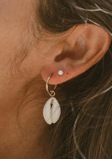 Close-up of an ear wearing a gold hoop earring with a shell pendant and a pearl stud.