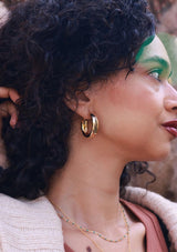 Woman with curly hair wearing gold hoop earrings and a necklace, outdoors.