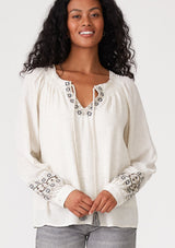 [Color: Natural] A front facing image of a brunette model wearing a bohemian blouse with embroidered details. With voluminous long sleeves, a split v neckline with tassel ties, a smocked neckline, and a relaxed fit. 
