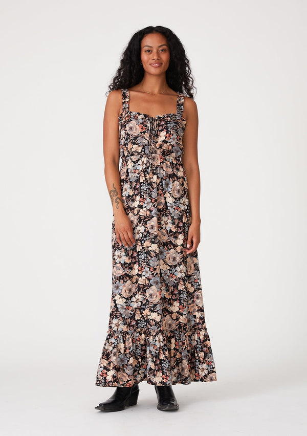 [Color: Black/Dusty Blue] A front facing image of a brunette model wearing a bohemian sleeveless maxi dress in a black and dusty blue floral print. With ruffled tank top straps, a tiered long skirt, an elastic waist, a square neckline, and a lace up tie front top. 