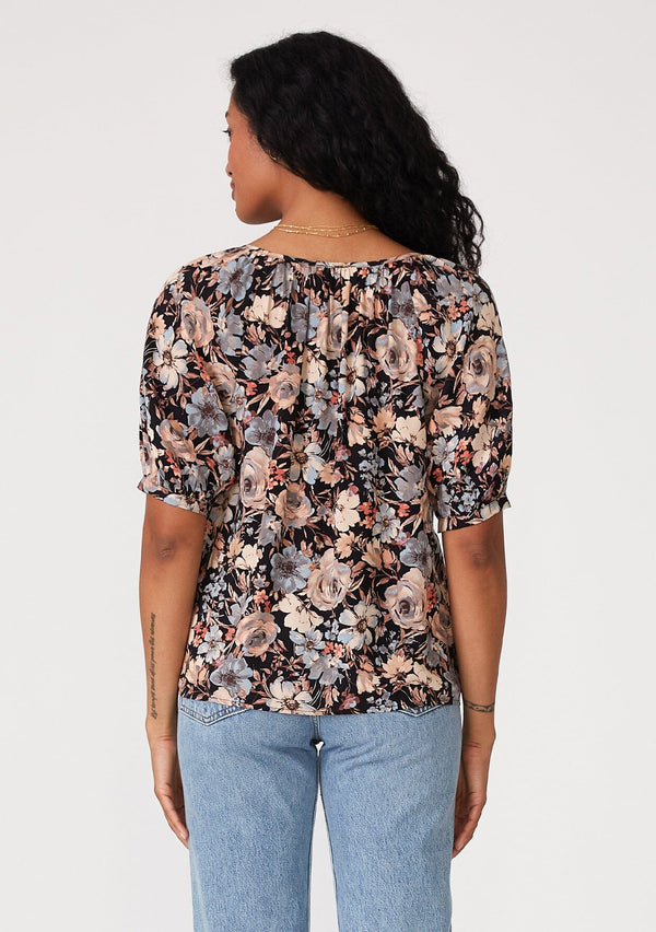 [Color: Black/Dusty Blue] A back facing image of a brunette model wearing a short puff sleeve blouse in a black and dusty blue floral print. With a split v neckline with tassel ties and a self covered button front. 