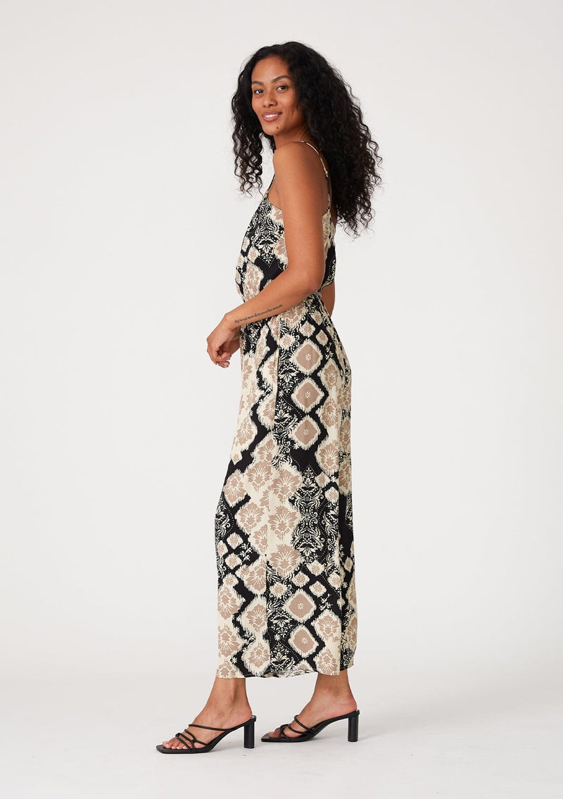 [Color: Black/Khaki] A side facing image of a brunette model wearing a sleeveless jumpsuit in a black and beige bohemian print. With adjustable spaghetti straps, a scoop neckline, side pockets, a long wide leg, and a beaded braided belt. 