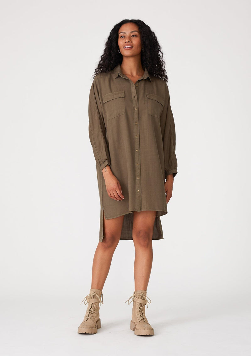 [Color: Military] A front facing image of a brunette model wearing an olive green relaxed fit shirt dress. With long sleeves, a button front, a high low hemline, a collared neckline, and front flap pockets. 