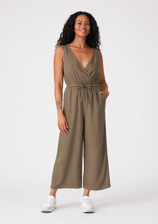 [Color: Military] A front facing image of a brunette model wearing an olive green sleeveless jumpsuit with a cropped wide leg, a surplice v neckline, side pockets, and a drawstring tie waist. 