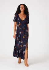 [Color: Navy/Mustard] A front facing image of a brunette model wearing a bohemian fall maxi dress in a navy blue and mustard yellow floral print. With short puff sleeves, a v neckline, a long flowy tiered skirt, and a side slit.