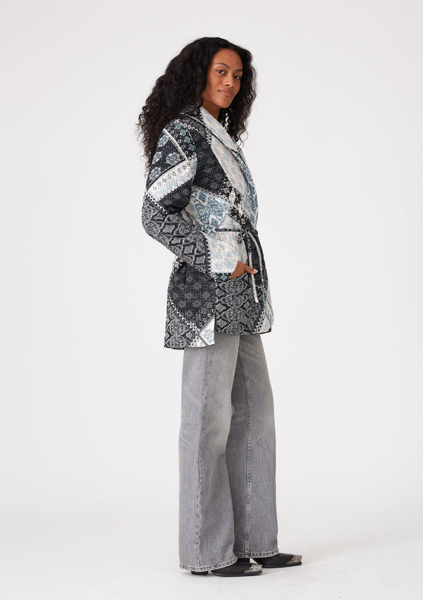[Color: Natural/Blue] A side facing image of a brunette model wearing a blue and white patchwork print quilted jacket. With long sleeves, a shawl collar, side pockets, and a tie waist belt. 