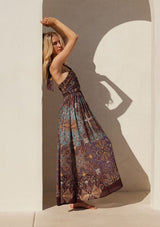 [Color: Raisin/Teal] A full body side facing image of a blonde model standing outside wearing a bohemian halter maxi dress in a multi colored purple and teal floral print. With a tie neckline, a slim fit bodice with a smocked elastic back, and a long flowy skirt.