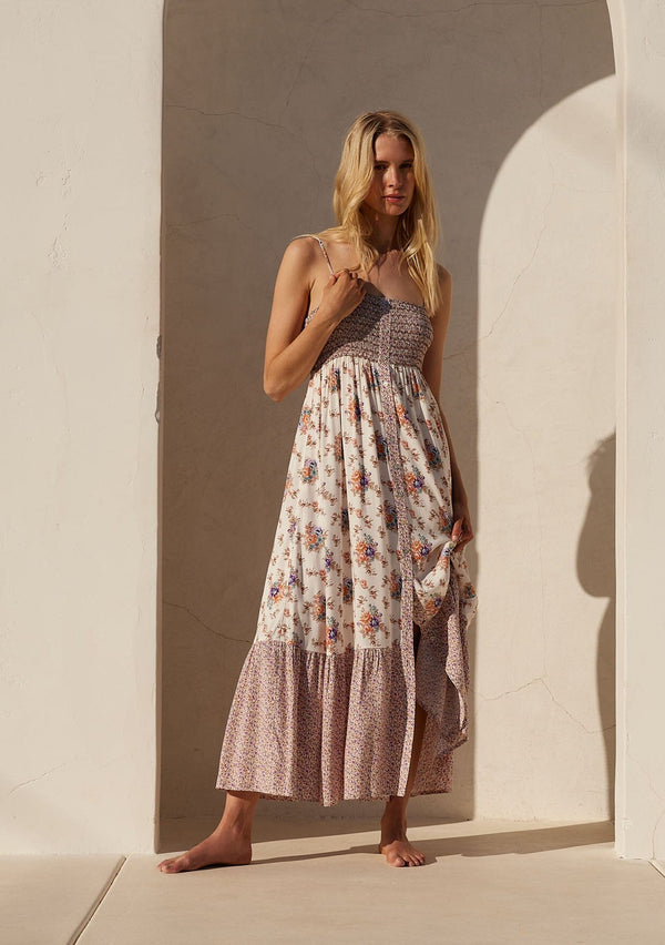 [Color: Natural/Purple] A front facing image of a blonde model standing outside wearing a best selling bohemian maxi dress in an off white and purple floral border print. With adjustable spaghetti straps, a square neckline, a smocked fitted bodice, a flowy tiered skirt, side pockets, and a button front.
