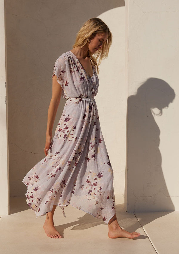 [Color: Dusty Blush/Wine] A side facing image of a blonde model standing outside wearing a bohemian maxi dress designed in a pink floral print and crafted from textured chiffon. With short dolman sleeves, a surplice v neckline, an open back with tie closure, and a flowy asymmetric hemline skirt.